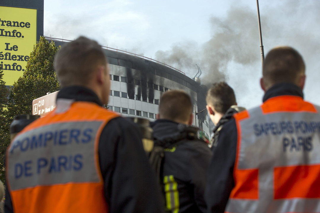 Bomberos observan una columna de humo se eleva en el aire desde uno de los últimos pisos del edificio de la radio pública francesa en París 