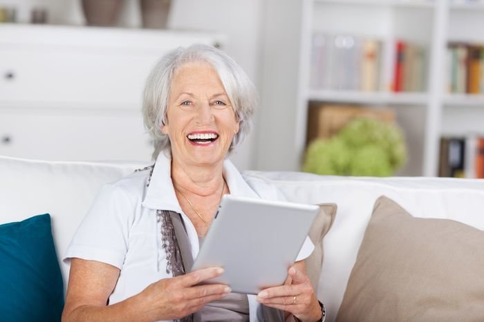 Laughing beautiful senior woman with a tablet-pc in her hands sitting on a sofa at home looking at the camera