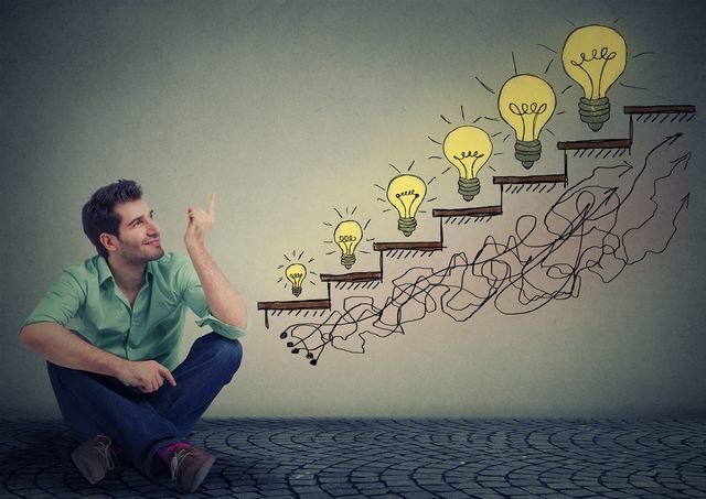 Happy man sitting on floor in his office pointing at business education success, promotion, company growth isolated gray wall texture background. Handsome guy looking up at growing up light bulbs