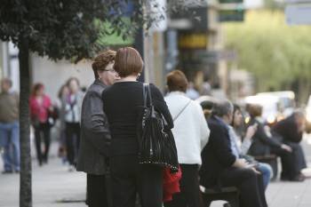 Dos mujeres conversan en la calle, junto a varios grupos de mayores sentados en los bancos. 
