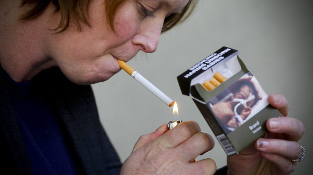 Una joven enciende un cigarrillo y muestra su paquete con una de las advertencias sanitarias.