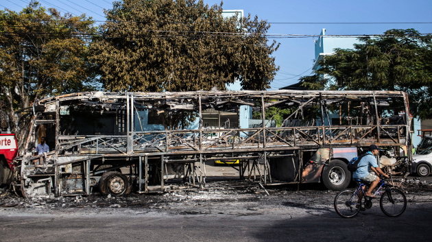Un hombre pasa en bicicleta frente a los restos de un autobús incendiado 