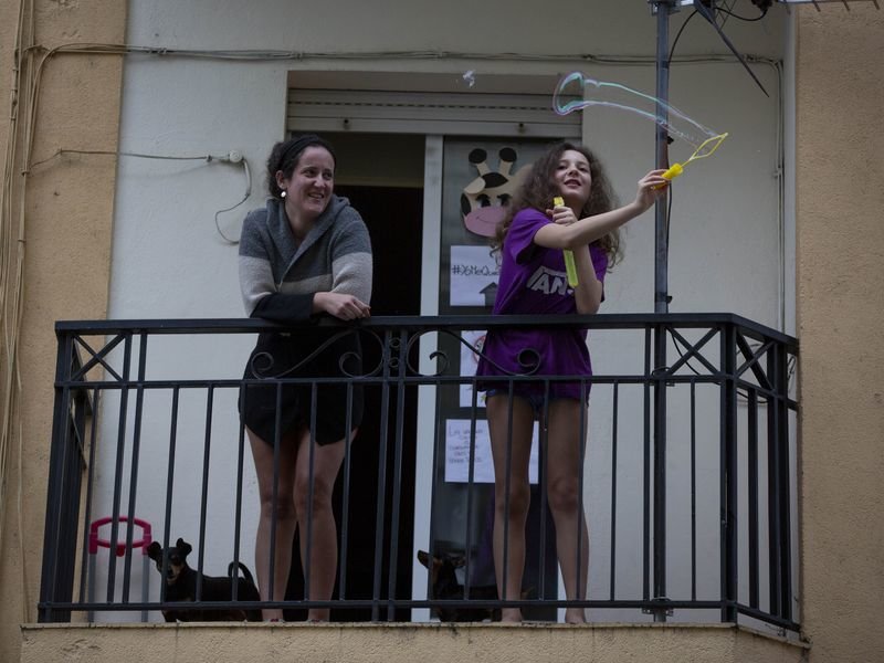 Una niña hace una pompa de jabón desde el balcón de su casa, acompañada de una familiar, en la ciudad (XESÚS FARIÑAS).