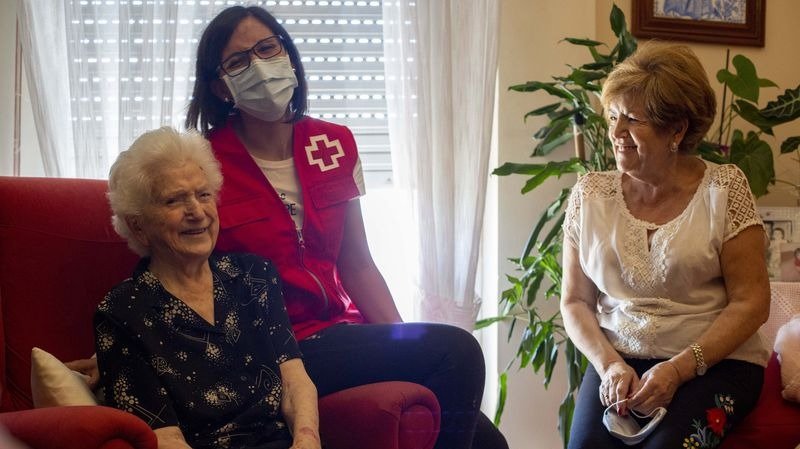 Celia Paz sonríe en el sofá de su casa, en la ciudad, observada por su hija Carmen. En el medio, les acompaña Natividad Álvarez, de Cruz Roja  (XESÚS FARIÑAS).
