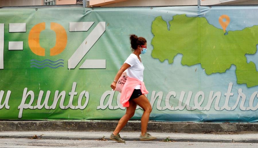 Una mujer con mascarilla camina frente a un cartel publicitario del pueblo de A Mariña. EFE/ Eliseo Trigo