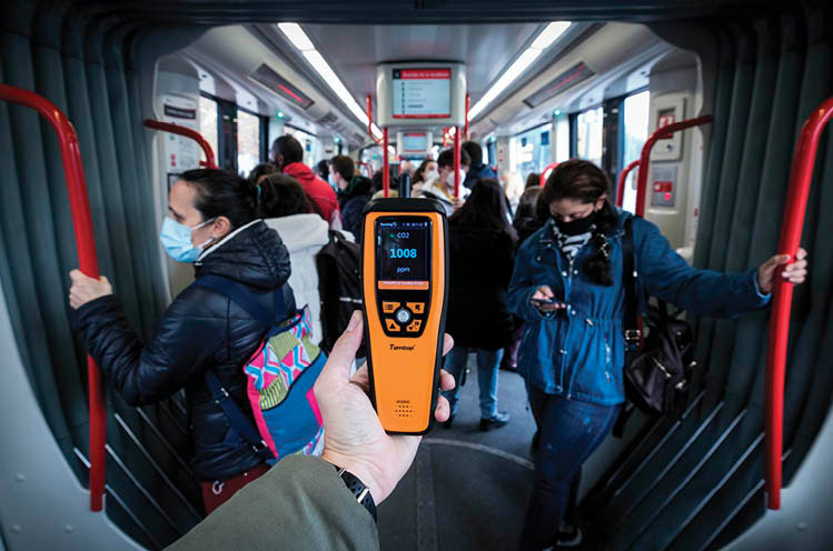 <p> Una mujer sostiene un medidor de dióxido de carbono en el interior de un autobús repleto de personas. </p>
